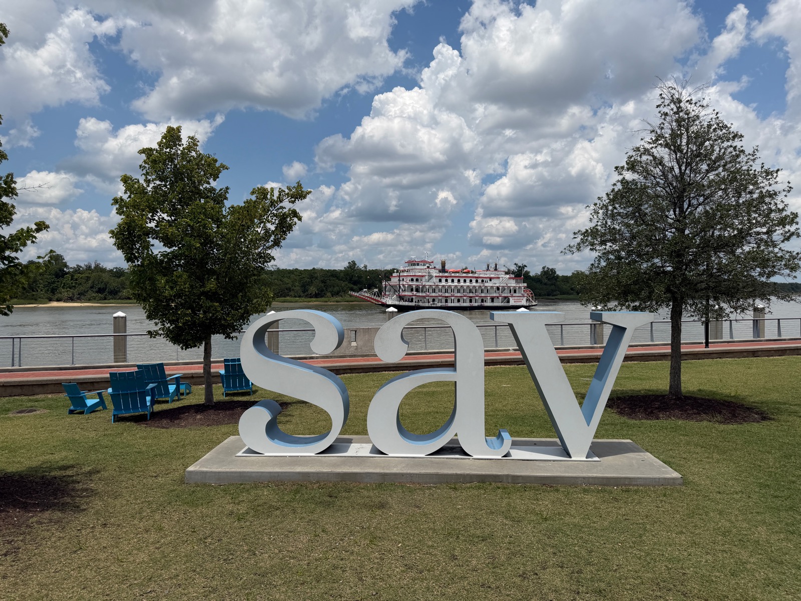 The white “sav” letter sculpture on Hutchinson Island’s riverfront lawn, the red-and-white Georgia Queen riverboat passing on the Savannah River behind it.